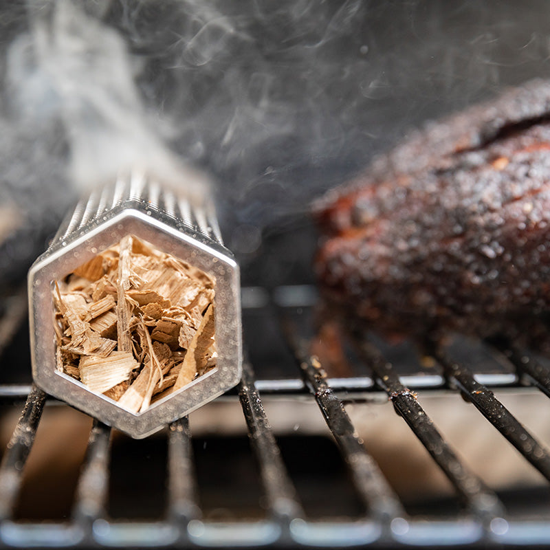 Smoker tube filled with smoking wood chips next to smoked BBQ meat on a grill
